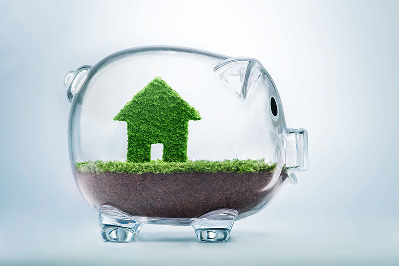 A transparent piggy bank contains soil and a small green house figure made of grass, symbolizing saving money for a home or eco-friendly housing investments.